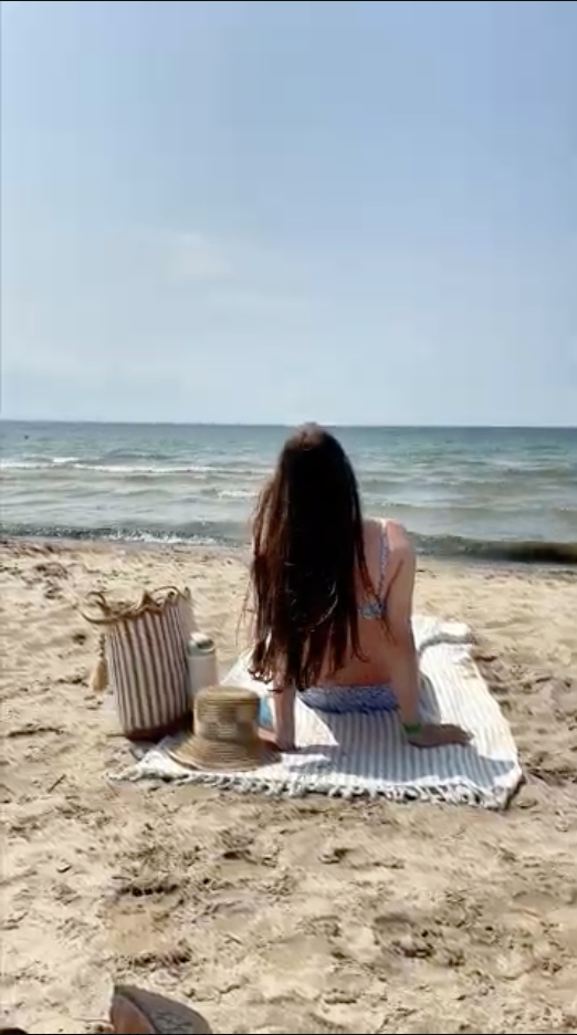 A woman sitting on a striped towel on the sand at Crystal Beach, looking out at Lake Erie, a straw hat and beach bag beside her.