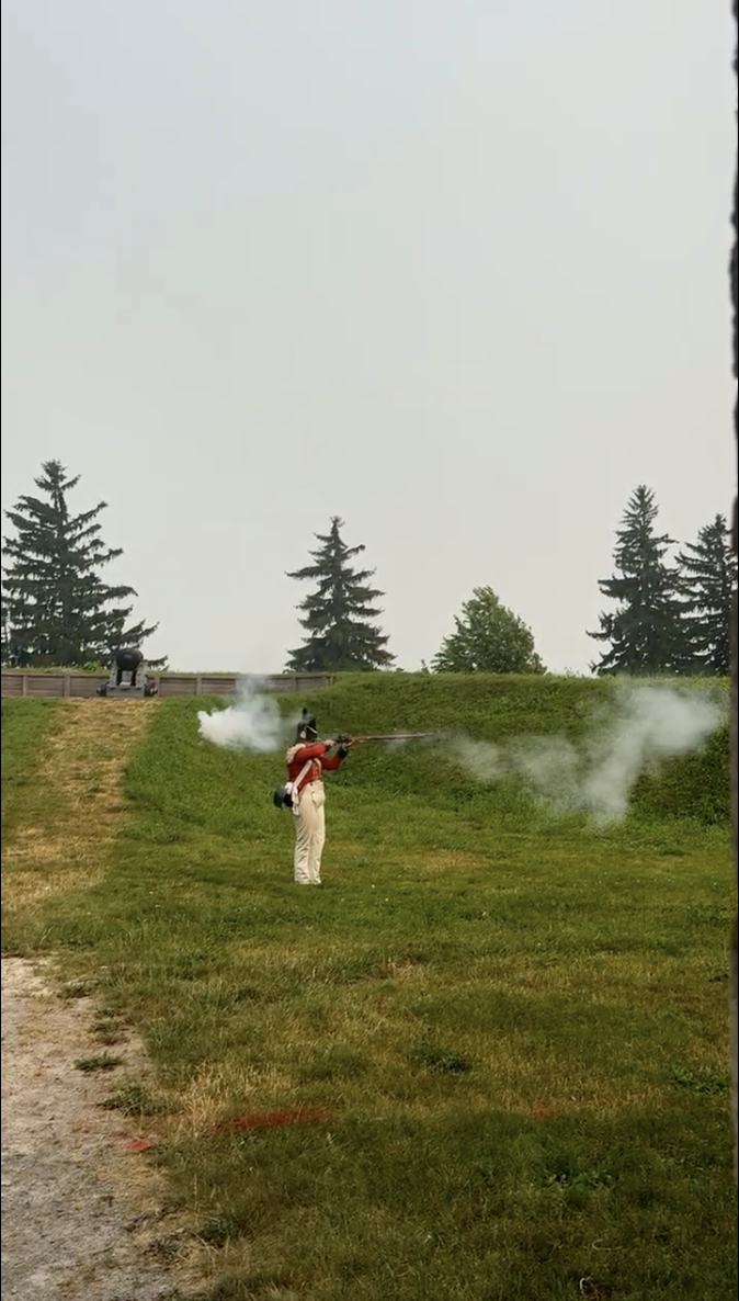 A costumed British soldier in red coat firing a musket on the grass earthworks at Old Fort Erie, smoke drifting across the rampart with a cannon behind.