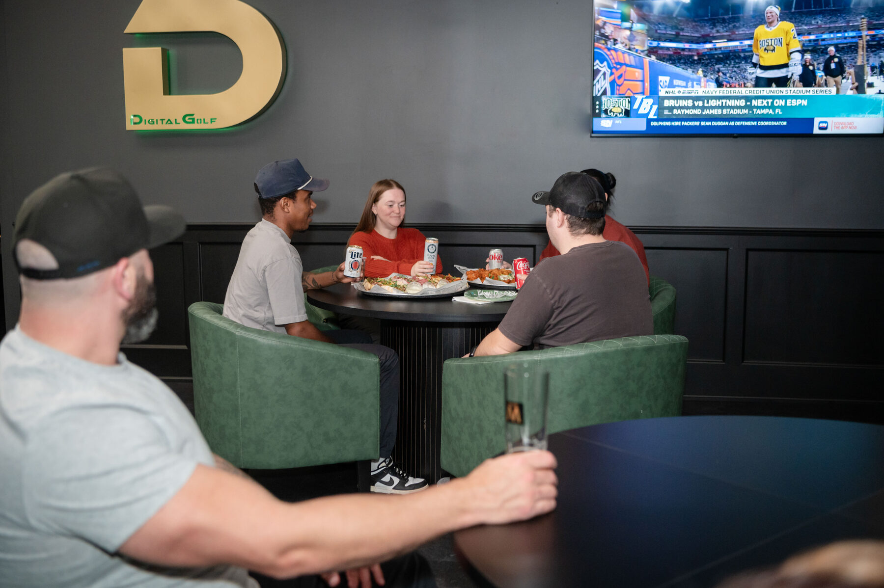 Customers with food and drinks in the lounge under the Digital Golf logo, sports on the TV.