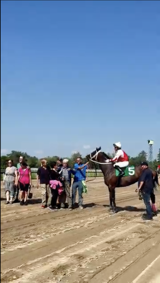 The winner’s circle at Fort Erie Race Track, a jockey in red and white silks on horse number 5 with the connections lined up on the dirt beside them.
