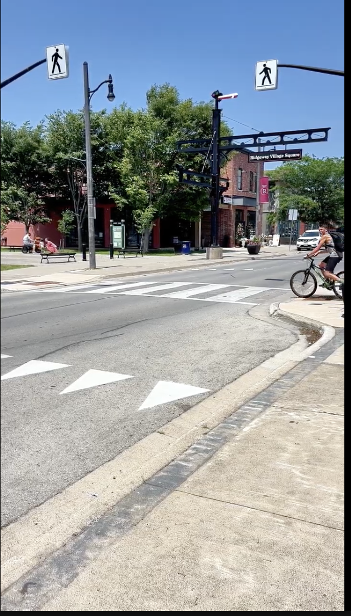 A cyclist crossing at Ridgeway Village Square, the black gateway arch and brick storefronts of the main street behind.