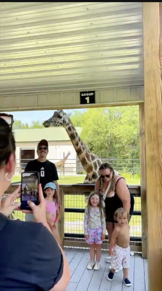 A family posing for a photo on the giraffe feeding deck at Safari Niagara, a giraffe leaning in over the rail beside them.